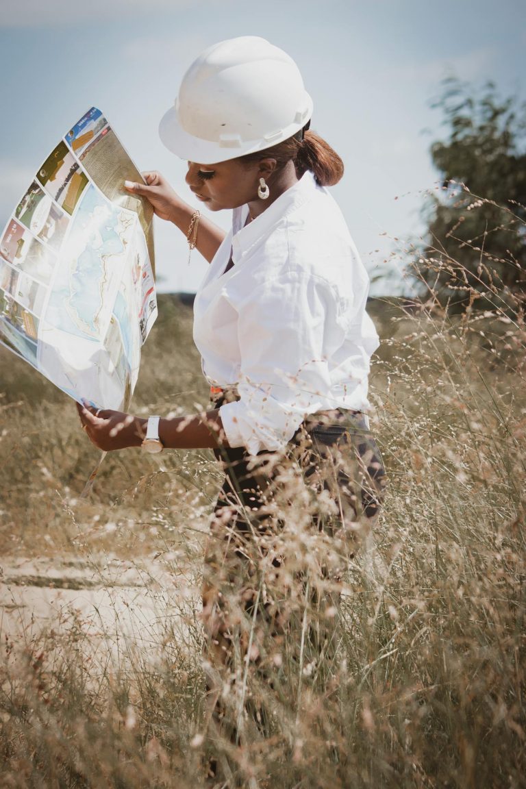 An African female engineer wearing a hard hat studies site plans in a rural setting, illustrating fieldwork.