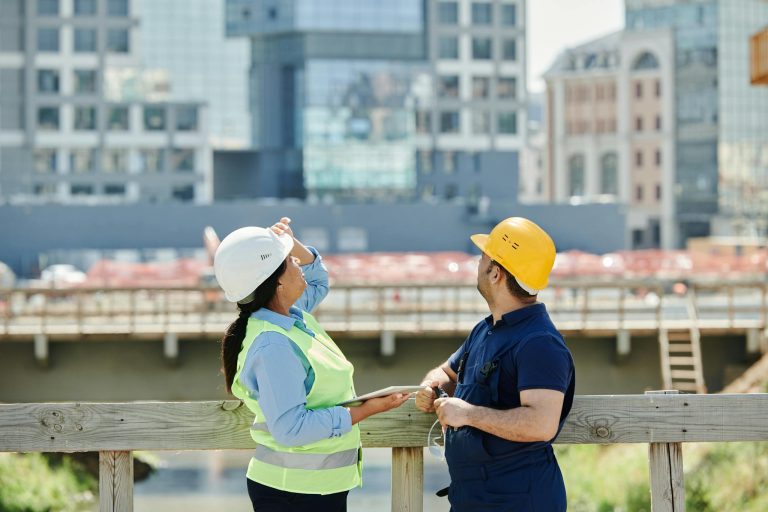 Engineers at a construction site reviewing architectural plans with modern cityscape background.